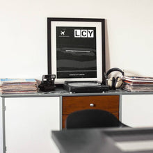 Lade das Bild in den Galerie-Viewer, London City Airport (Flughafen LCY / EGLC) Dark Poster - Minimal - Vorschau auf einem Tisch mit Plattenspieler und Kamera
